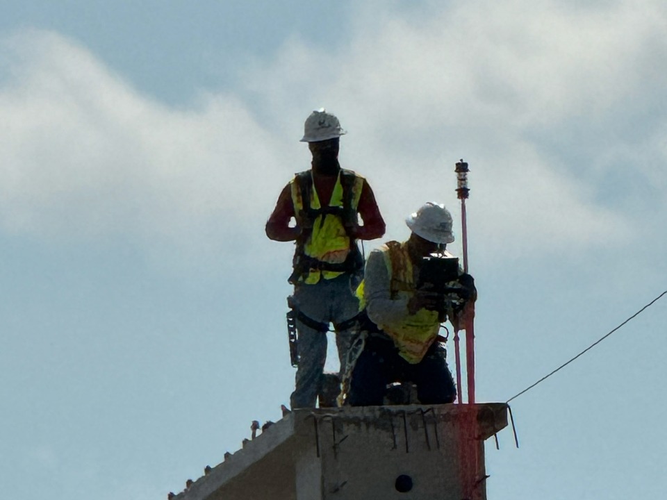 Two construction workers on a bridge deck under construction, one holding a survey prism pole
